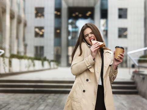 Young beautiful businesslady holding planner under arm walking downtown near office centers with cup of coffee bites sandwich in a hurry, multitasking concept