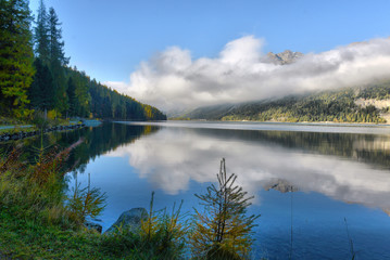 Fototapeta premium Mountain lake at sunrise in autumn. Landscape with lake, gold sunlight, blue fog over the water, reflection, trees with colorful leaves