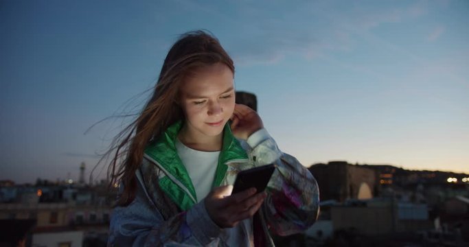 Portrait of nice pleasant Caucasian female staying up to date in social app outdoor, evening windy moment at terrace of happy cute girl influencer using smartphone online in internet connected