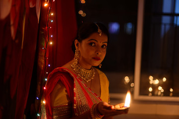 Fototapeta na wymiar An young and beautiful Indian Bengali woman in Indian traditional dress is sitting while holding Diwali diya/lamp in her hand in front of colorful bokeh lights. Indian lifestyle and Diwali celebration