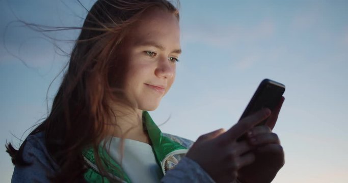 Close up portrait of pleasant pretty Caucasian scrolling application newsfeed outdoor, smiling young student teen girl with smartphone evening sky on background wind gently touching ginger hair