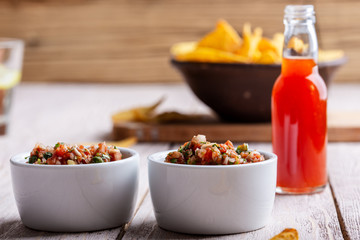 Two bowls full of salsa dip, hot red chili sauce bottle and tortilla chips