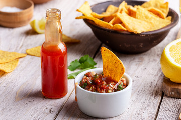 Two bowls full of salsa dip, hot red chili sauce bottle and tortilla chips