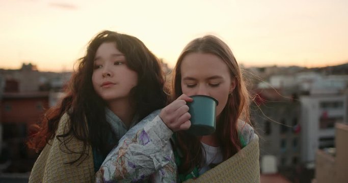Portrait of two charming mixed race female friends wrapped in cozy plaid together outdoor, serene dreamy young teen girls drinking tea from metal cup at rooftop enjoying lovely evening city view