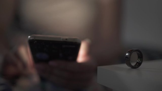 White woman with brunette hair using her phone before putting a smart ring on finger and going to sleep.