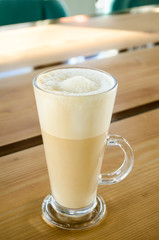 Tall transparent glass beaker with a handle, with foam sugar latte inside, on a wooden table in a cafe in the sunshine