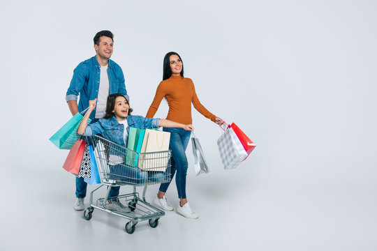 Having fun. Full-length photo of three happy family members, who are enjoying their time together in a shopping center, carrying a lot of multicolored paper bags and riding a small child in a cart.