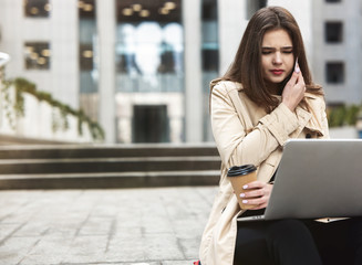 Obraz premium young beautiful businesswoman works in her laptop drinks coffee sitting on stairs near office center in downtown, looks upset, multitasking concept
