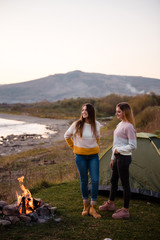 Two young women camping. Friends travel and relax comfortably in the open air by the fire. Camping on green meadow on river and mountain background