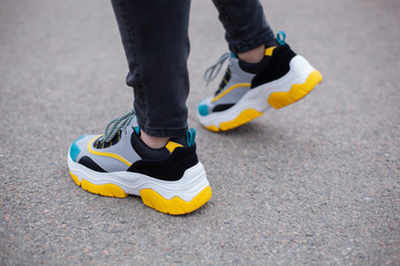 Feet shod in sneakers multi-colored yellow, white, black and blue are on the asphalt road. Close-up