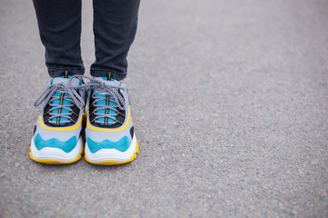 Feet shod in sneakers multi-colored yellow, white, black and blue are on the asphalt road. Close-up