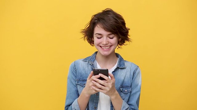 Smiling young woman in denim jacket white t-shirt posing isolated on yellow background in studio. People sincere emotions, lifestyle concept. Looking at cellphone using mobile phone chatting have fun