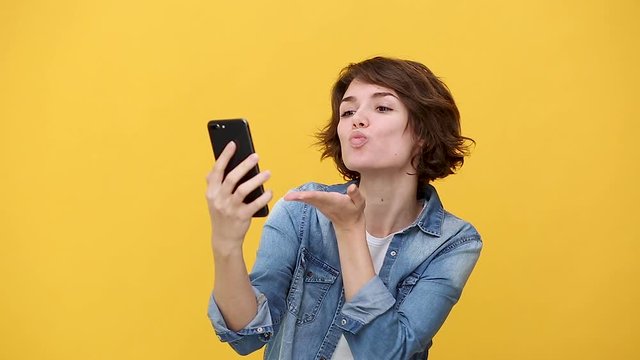 Pretty young woman in denim jacket white t-shirt posing isolated on yellow background in studio. People lifestyle concept. Looking at photo camera with charming smile doing selfie shot on mobile phone