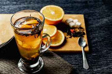  Citrus drink, tea with petals in a transparent glass mug with slices of sweet orange and refined sugar. Slicing fruit on a wooden Board with a brown napkin in a cozy dark background in a low key