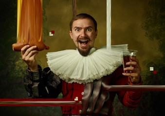 Hot dog and beer. Young man as a medieval knight on dark studio background. Portrait of man in retro costume. Emotions, comparison of eras. Stylish creative design, art vision, new look of artwork.