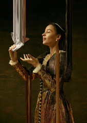 Fototapeta premium Medieval young woman in brown vintage clothing as a duchess on dark studio background. Trying chocolate, sweets. Concept of comparison of eras. Stylish creative design, art vision, new look of artwork