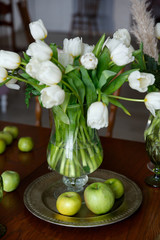 white tulips in a glass vase and green apples on a plate on the table