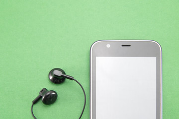 Black smartphone with a white border (stripe) in a circle. Gray blank screen. and two black small earbuds from the headset for insertion into the ear. Part of the phone on a green isolated background