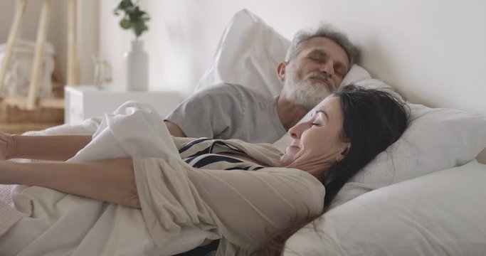 Side view close-up of Caucasian woman stretching in the morning in bed, sleeping husband hugging lovely wife. Happy senior family resting on weekends at home. Love, lifestyle, joy. Cinema 4k ProRes HQ
