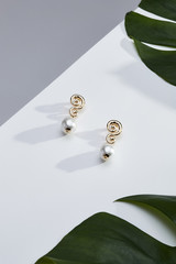 Subject shot of a pair of stud earrings on the white and gray surface surrounded with green tropic leaves. Each earring is made as a golden tylized infinity sign with a pearl bead pendant.