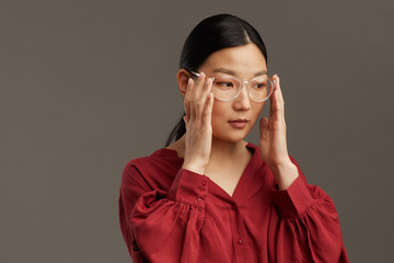 Head and shoulders portrait of elegant Asian woman putting on glasses while standing against grey...