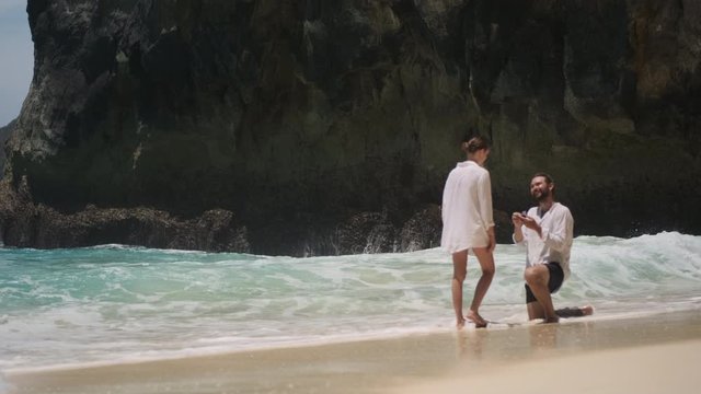 Young handsome man doing proposal to his gorgeous girlfriend on empty tropical beach of paradise island. Touching moment of couple getting engaged