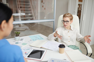 Portrait of modern short haired woman wearing glasses arguing with colleague or partner across...