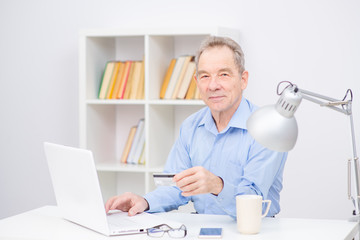 Obraz premium An elderly man looking at camera with a credit card in his hand. Senior citizen working at a laptop with a credit card in his hands
