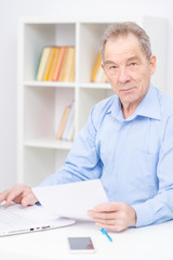 Senior sitting near a laptop, holding papers in his hand and looking at camera.