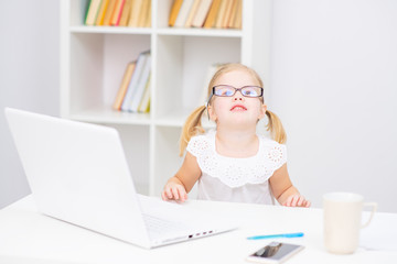 Little girl wearing glasses in a white blouse sitting near a laptop and looking at the camera with a smile