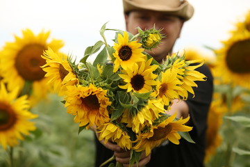  young man in a beige hat and a black t-shirt in a field where sunflowers with a bouquet enjoy the summer landscape