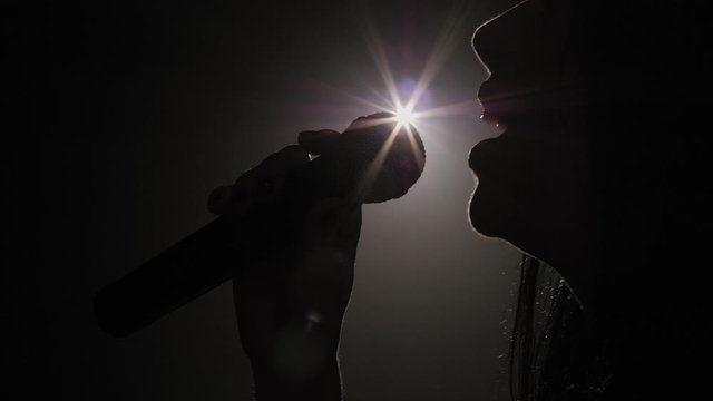 Silhouette of a luxurious singer in an evening gown on a stage in the dark. The girl sings a song on an empty stage in the light of a white spotlight.