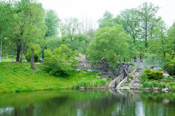 Fototapeta na wymiar Scenic park landscape with stone bridge and wheel of dam near pond with reflections of green trees and plants.