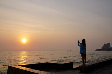 Fototapeta premium silhouette of woman take photo sunset on the sea and colorful sky by smartphone.