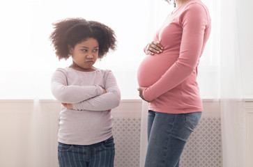 Offended little black daughter looking at pregnant mom's belly with jealousy