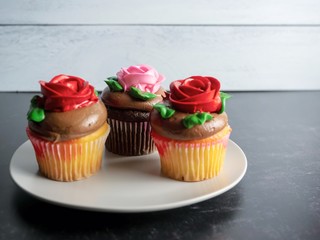 3 cupcakes sitting on a plate, yellow and chocolate with chocolate frosting and red and pink roses made from frosting, on a black slate surface with a gray shiplap background, festive and delicious.