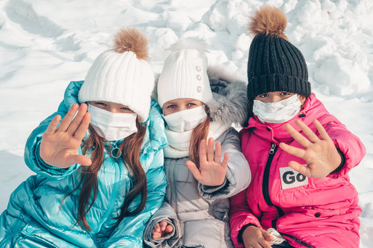 Three teenage girls in protective masks are sitting on the snow in the park. Girls show a gesture of denial. Stop the epidemic of viruses and flu. Stop coronovirus