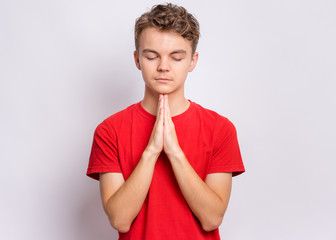 Portrait of teen boy praying, on grey background. Cute caucasian teenager with hands folded in...