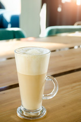 Tall transparent glass beaker with a handle, with foam sugar latte inside, on a wooden table in a cafe in the sunshine