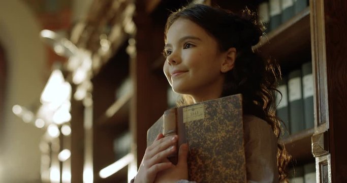 Close up of small teen Caucasian smart girl reading old book in dark library with magical atmosphere. Cute kid dreaming about fairytale and smiling happily in bibliotheca passage.