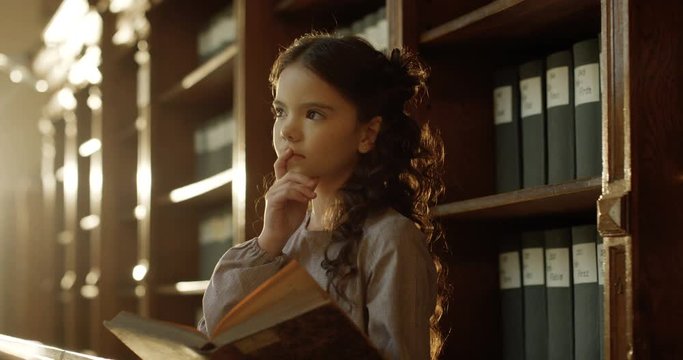 Little beautiful Caucasian girl reading book of fairytales in dark library and dreaming. Cute kid smiling happily while flipping pages and watching pictures of tales in bibliotheca.