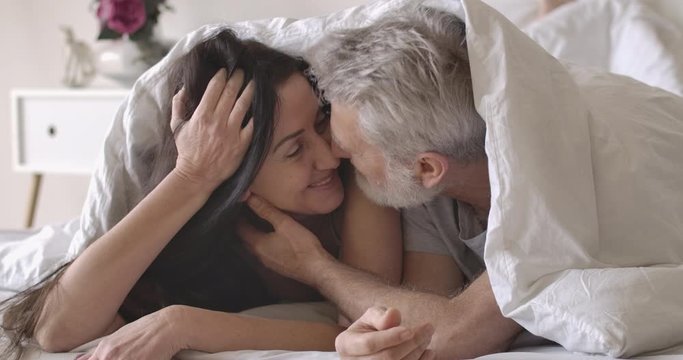Close-up portrait of smiling senior Caucasian couple lying under white blanket and rubbing noses. Happy husband touching wife's face in the morning in bed. Love, romance, lifestyle. Cinema4k ProRes HQ