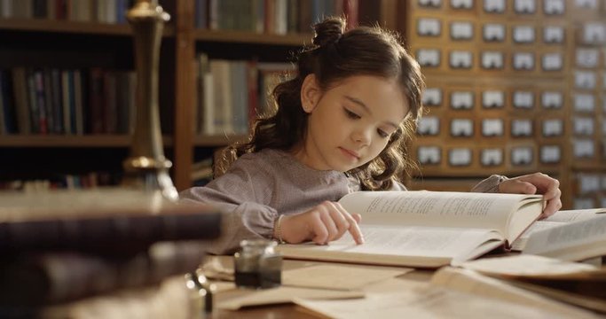 Caucasian small pretty and cute girl sitting at desk in library and reading bok attentively. Little beautiful kid studying old textbook at table in bibliotheca.