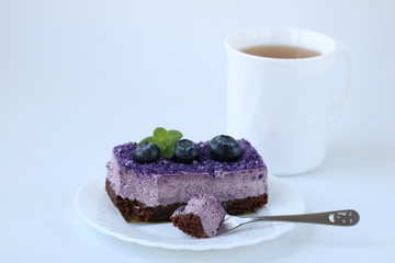 bitten off tasty purple blueberry mousse cake with blueberry on white plate with teaspoon and cup of tea on white background, horizontal, side view, copy space, selective focus.