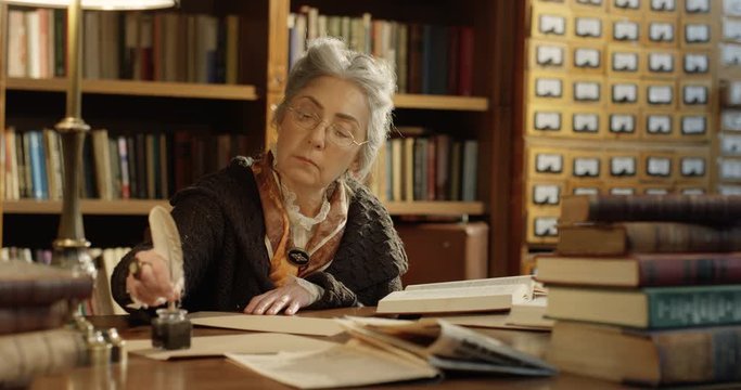 Caucasian senior woman with big ring on finger writing letter with feather and composing text. Wise female old writer with plume sitting at table in library and using ink and pot. Antique concept.