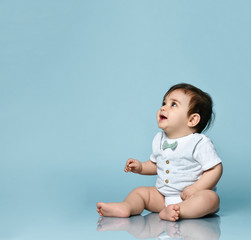 Little toddler in white bodysuit as a vest with bow-tie, barefoot. He is sitting on the floor against blue background. Close up