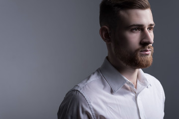Fototapeta premium Close-up, studio dramatic portrait of a handsome bearded guy in a white shirt. On a gray background.
