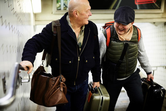 Smiling senior gay couple with luggage walking on staircase in subway