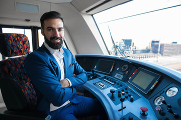 Train driver occupation. Portrait of young bearded professional driver in uniform with crossed arms sitting in train cockpit.