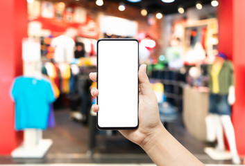 Hand holding smartphone in clothing store. Mock up mobile phone image of hand holding smart phone with blank white screen for your advertisement on blurred background of clothing shop.